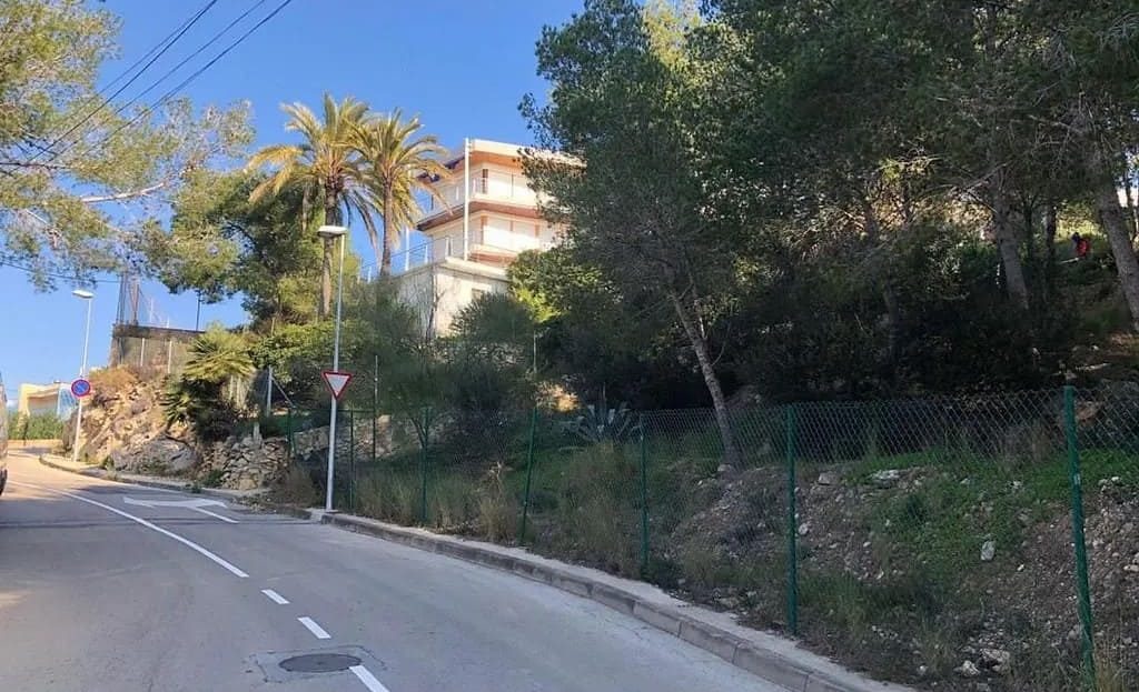 Уличный вид на подъездную дорогу к участку (Carrer del Cap de Salou). Показана ухоженная асфальтированная дорога с разметкой, тротуаром и существующими жилыми домами на заднем плане, что подтверждает наличие городской инфраструктуры рядом с землей